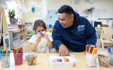 this is a photo of a little girl holding up a blue marker to her face, in the midst of coloring at a table with her dad smiling and sitting next to her.