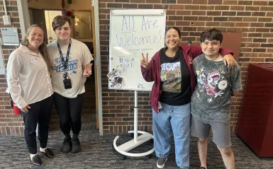 this is a photo of two female NHCC Diversity Specialists standing and smiling with two NHCC students in front of the Gerry Huerth Center in the Campus Center. The whiteboard sign in between them reads, All Are Welcome! 