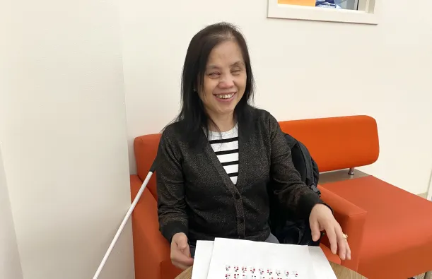 This is a photo of NHCC Student, May Spooner, smiling while sitting in the library on a comfortable chair with her cane at her side and a worksheet of the braille alphabet on the table in front of her along with a braille worksheet on Chapter 4 of the Harriet Tubman textbook she’s reading for her EAP class. 