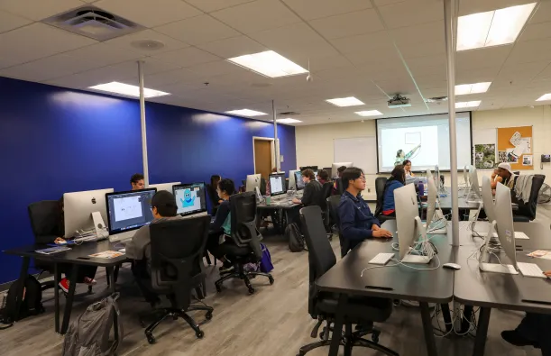 this is a photo of a graphic design classroom in NHCC's Fine Arts Center. Students are working at the iMac desktop computer stations, sitting in comfortable chairs, the teacher is pointing at a graphic on the screen at the front of the room, the wall is bright, dark blue and the vinyl floor is updated. 