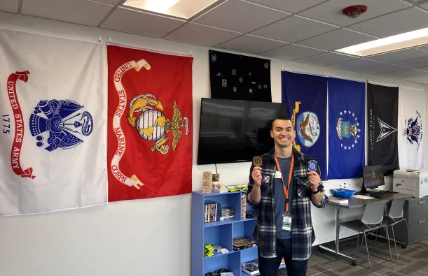this is a photo of NHCC staff, Dylan Rowinski holding up NHCC's student veteran patches and smiling in our Veterans Resource Center on campus. 