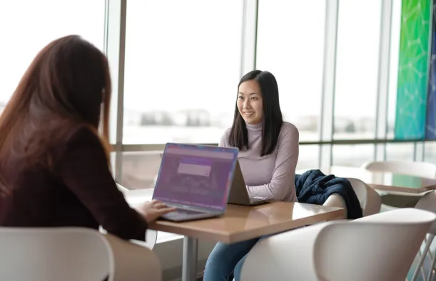 this is a photo of two female students studying at a table in the BHCC building, near a window, with their laptops on the table. One girl is smiling and looking at the camera and you can only see the back of the other girls head. 