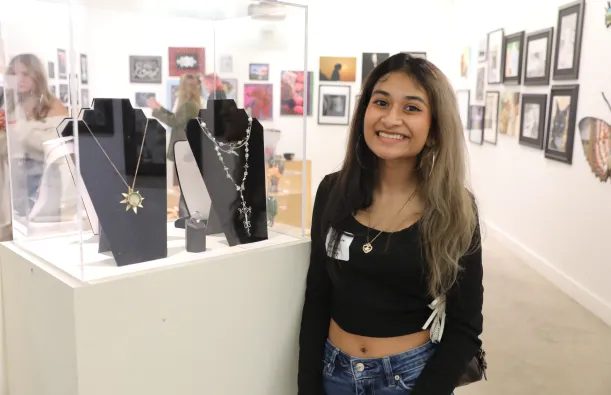 this is a smiling photo of a Maple Grove High School student named Willow Johri taken at the Northwestern High School Art Show Awards Ceremony evening. She is standing in NHCC's Art Gallery, next to a jewelry case with a gold sunshine necklace that she designed in it. 