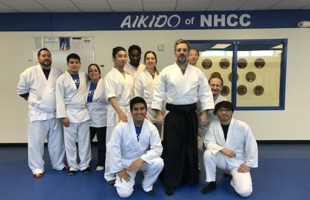 this is a group photo of NHCC instructor, Mark Larson standing with ten of his aikido students in the dojo on-campus, they are all wearing white robes and white pants, smiling for the photo. A sign in the background reads, 'Aikido at NHCC.' 