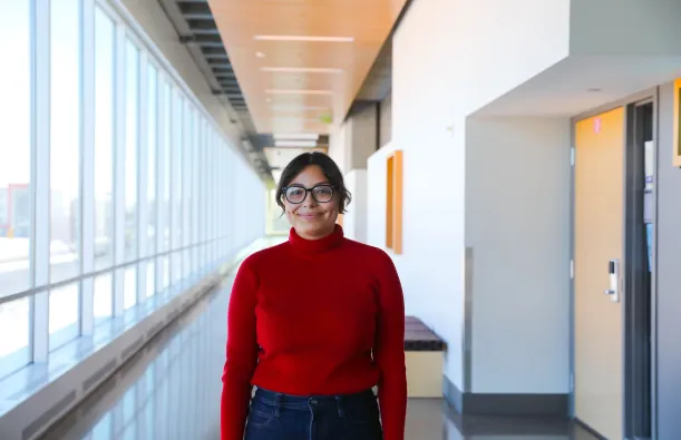 this is a photo of a female NHCC student smiling in the upstairs hallway of the BHCC building. She is wearing a red turtleneck sweater and dark wash jeans with her hair pulled back. 