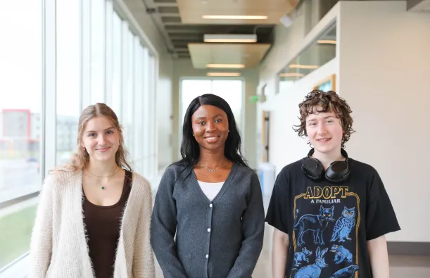 this is a photo of three NHCC students standing side-by-side in the upstairs hallway of the BHCC builidng. Two female students, Anas and Danista stand next to male student, Kenny, posing for a photo and smiling. 