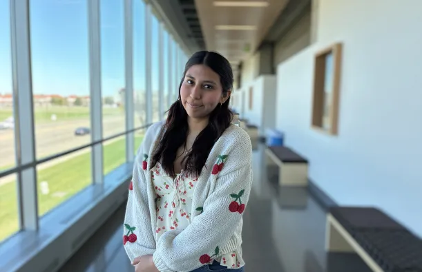 this is a photo of female NHCC student, Reyna Hernandez, standing in the upstairs hall of the BHCC building, smiling, wearing a tank top and a knit cardigan sweater that both have red cherries on them. 