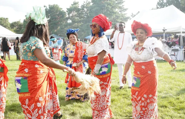 Several attendees wearing vibrant red, white, and multicolored traditional Igbo clothing and headwraps dance in a grassy outdoor festival area during the IgboFest Cultural Celebration. White event tents and other attendees are visible in the background.