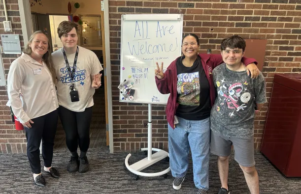 this is a photo of two female NHCC Diversity Specialists standing and smiling with two NHCC students in front of the Gerry Huerth Center in the Campus Center. The whiteboard sign in between them reads, All Are Welcome! 