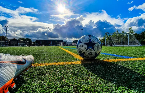 this is a stock image photo of a foot with a soccer cleat on, about to kick a soccer ball on a field on a bright sunny, blue sky with puffy white clouds day. 