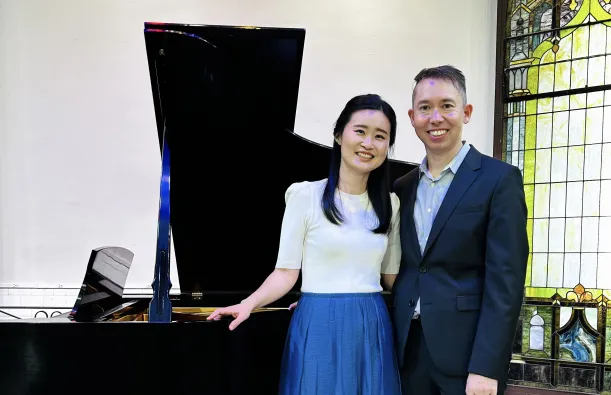 this is a smiling photo of NHCC music faculty, Grace Choi and Kevin Hobbs standing next to a piano. 