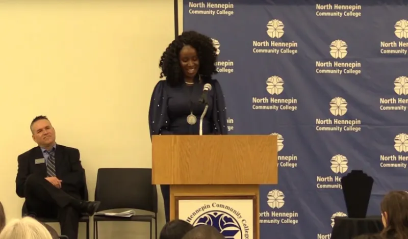 woman presenting on a stage in front of a podium man sitting behind her