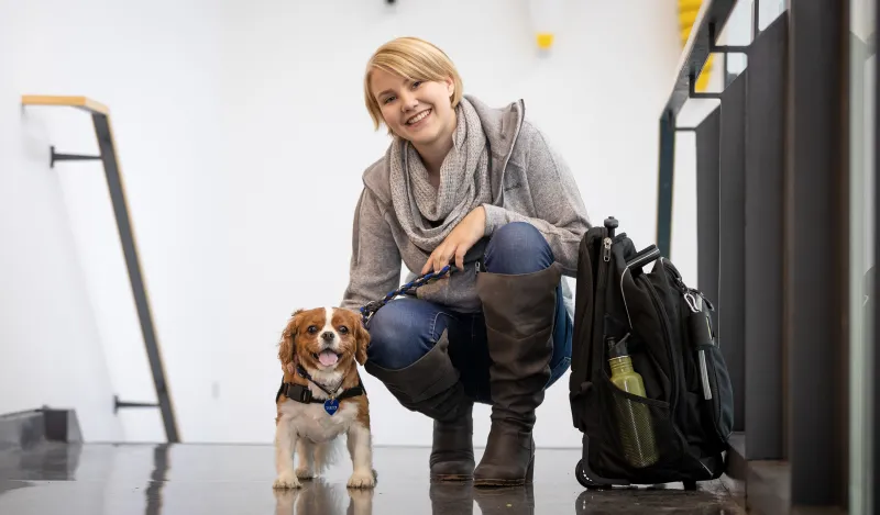 female student smiling and squatting next to a small service dog