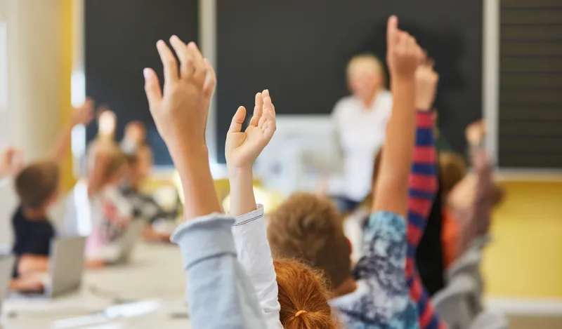 students raising their hands with a teacher in the background 