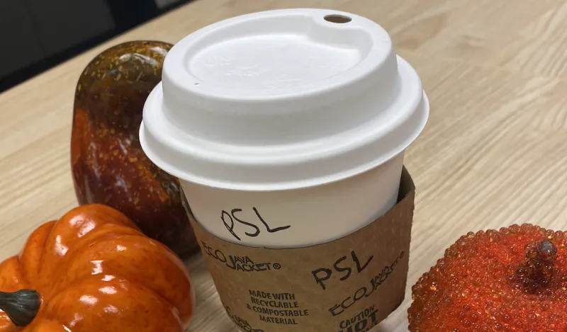 a white, paper coffee cup, with "PSL" written on it in Sharpie marker, next to two mini orange pumpkins 