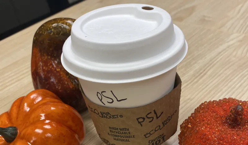 a white, paper coffee cup, with "PSL" written on it in Sharpie marker, next to two mini orange pumpkins