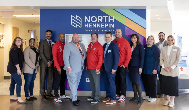 a group photo of staff and administrators from North Hennepin and St. Cloud State University posing for a photo in front of the NHCC sign in the ES building. 
