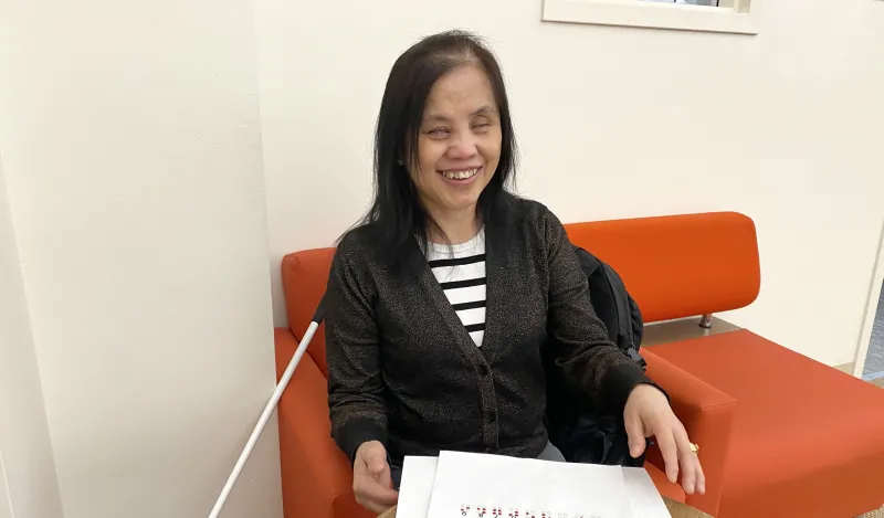This is a photo of NHCC Student, May Spooner, smiling while sitting in the library on a comfortable chair with her cane at her side and a worksheet of the braille alphabet on the table in front of her along with a braille worksheet on Chapter 4 of the Harriet Tubman textbook she’s reading for her EAP class. 