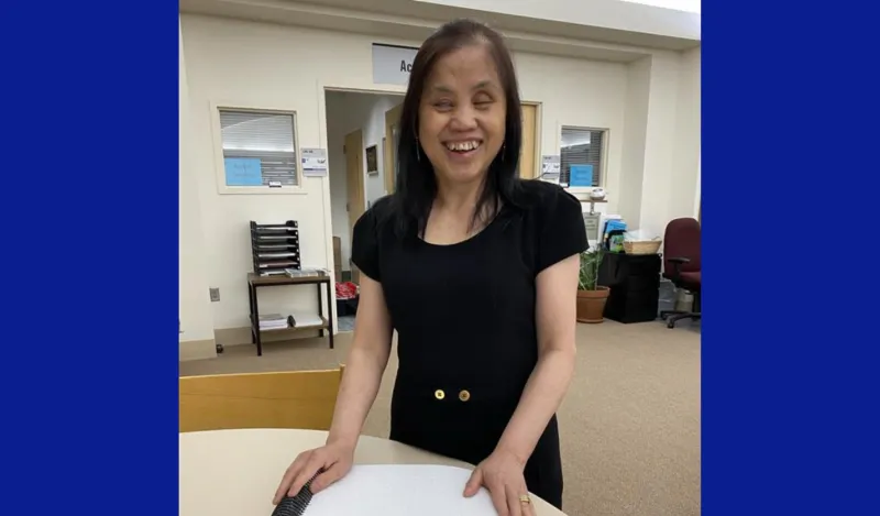 This is a photo of NHCC student, May in the library on-campus, outside of the Access Services offices, standing and smiling beside a table, hands on her newly printed braille textbook. 