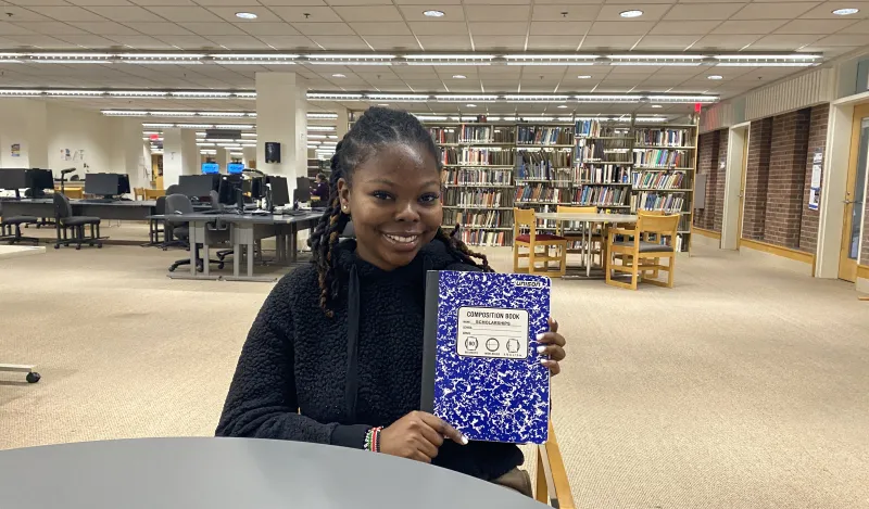 this is a photo of NHCC student, Lizzie, sitting in the library, posing with her blue composition notebook that she filled up with scholarships.