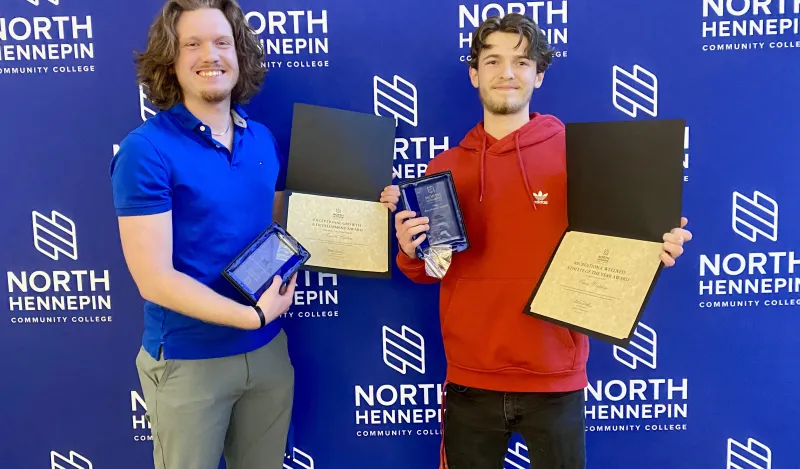 This is a photo of NHCC students and friends, Owen and Travis standing in front of an NHCC logo "step and repeat" backdrop smiling, both holding their certificates and awards, which are plaques made of glass, after being recognized at the Annual Student Life Awards Banquet. 