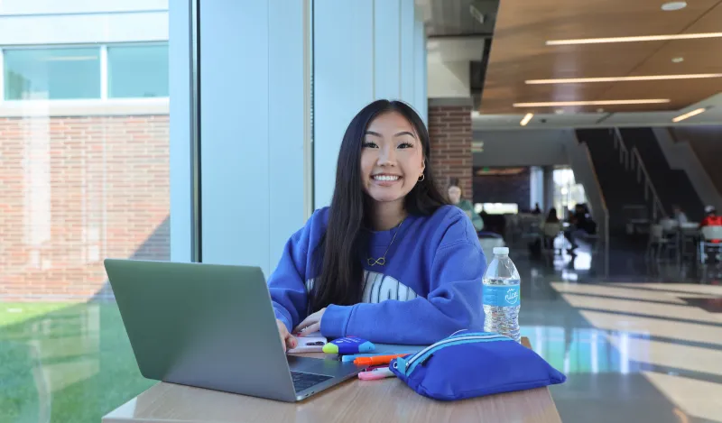 This is a candid photo of a female student smiling for the camera as she looks up from studying at a table in the BHCC hallway. The photo was taken in 2022.