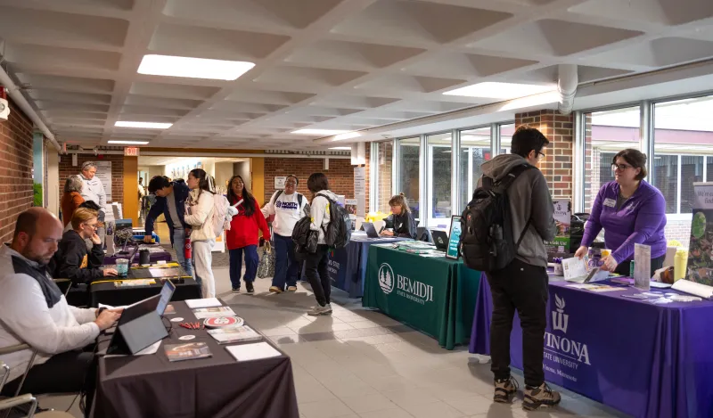 This is a photo of students walking through the ES and LRC hallway mingling with representatives from four-year colleges who are sitting there at tables in the hall to tell students about the transfer process. 