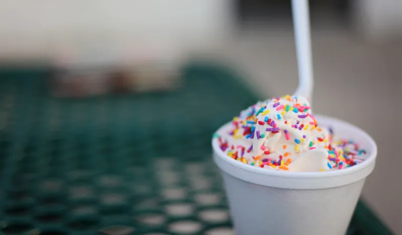 this is a photo of a cup of vanilla ice cream with rainbow sprinkles and a spoon sitting on a picnic table. 