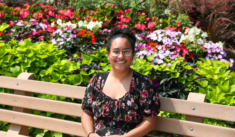 this is a photo of former NHCC student, Estefanny Zelaya, sitting outside on a bench, smiling, with colorful flowers surrounding her. 