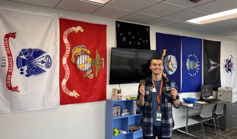 this is a photo of NHCC staff, Dylan Rowinski holding up NHCC's student veteran patches and smiling in our Veterans Resource Center on campus. 