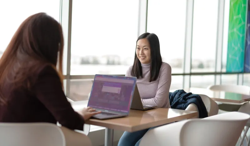 this is a photo of two female students studying at a table in the BHCC building, near a window, with their laptops on the table. One girl is smiling and looking at the camera and you can only see the back of the other girls head. 