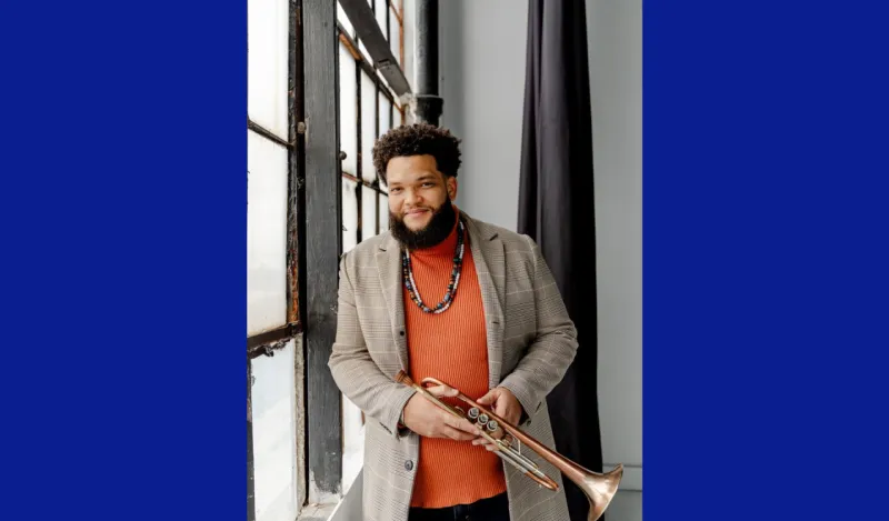 this is a photo of guest musician, Kris Johnson, standing next to a window, holding his trumpet. 