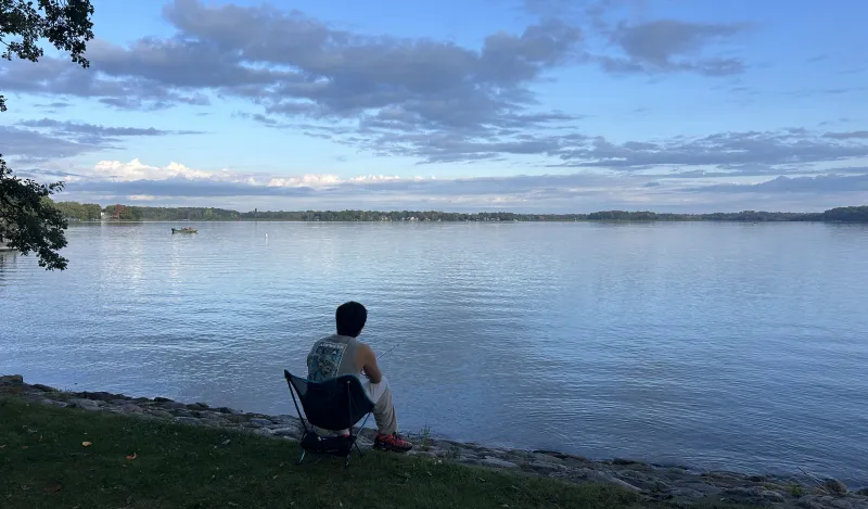 this is a photo of a person sitting by a lake with their back to the camera fishing. The sky and the lake are blue. 