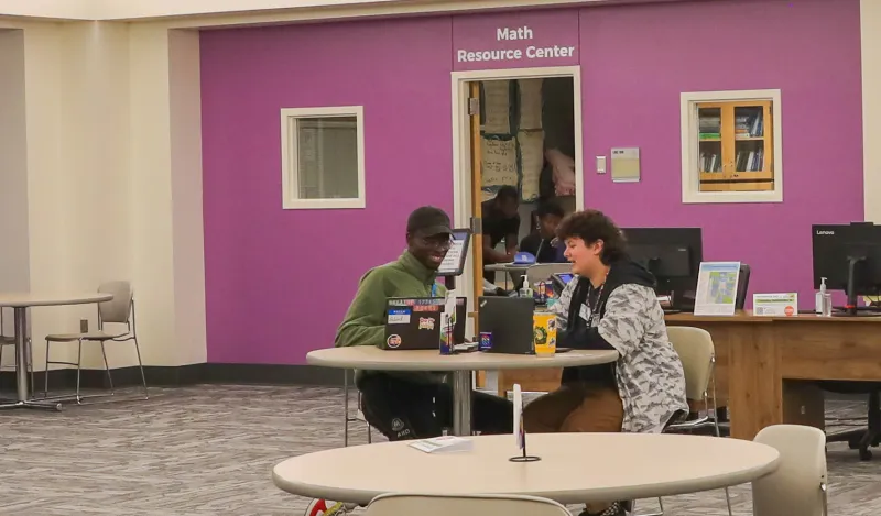 this is a photo of an NHCC tutoring employee sitting at a table working with a student. They are both looking at their open laptops, smiling, at a table in NHCC's Tutoring Center. A purple wall is behind them, with a sign that reads, 'Math Resource Center.'