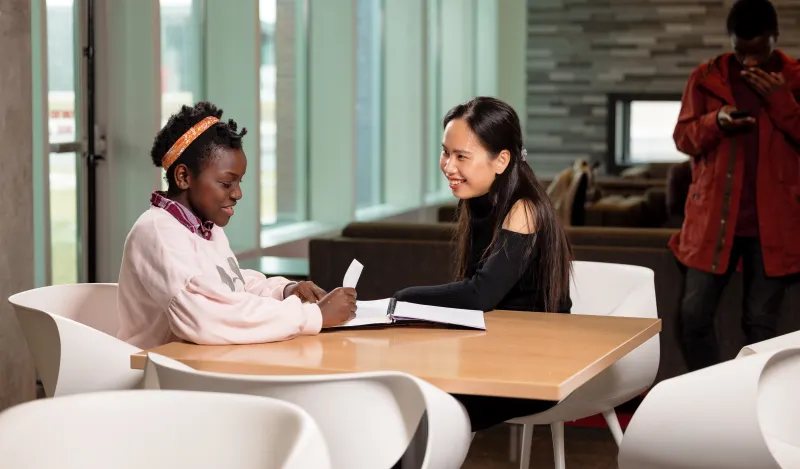 this is a photo of two female NHCC students sitting in chairs at a table by the coffee shop on-campus. The fireplace is in the background, and they are smiling at each other, while looking at papers in a notebook on the table. 