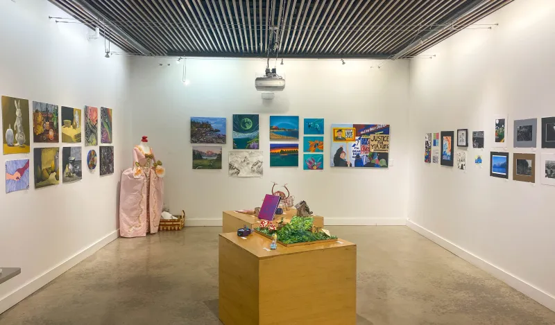 this is a photo of an empty NHCC Art Gallery, with art on the walls and tables, the lights are on and there's a 3D sculpture in the corner from the spring 2025 NHCC Student Art Show. 