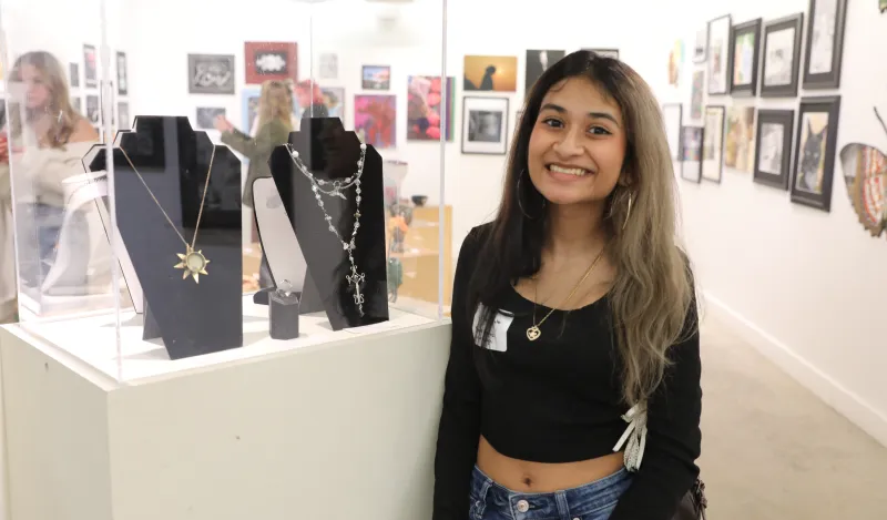 this is a smiling photo of a Maple Grove High School student named Willow Johri taken at the Northwestern High School Art Show Awards Ceremony evening. She is standing in NHCC's Art Gallery, next to a jewelry case with a gold sunshine necklace that she designed in it. 