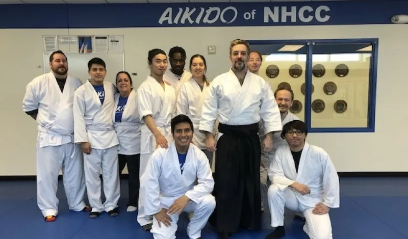 this is a group photo of NHCC instructor, Mark Larson standing with ten of his aikido students in the dojo on-campus, they are all wearing white robes and white pants, smiling for the photo. A sign in the background reads, 'Aikido at NHCC.' 
