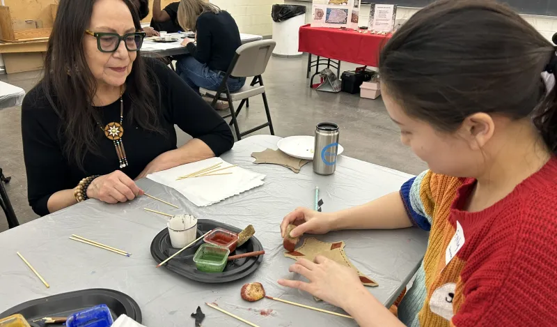 Artist Fern Cloud with a student using a traditional bone brush and natural pigments to paint a sample animal hide.