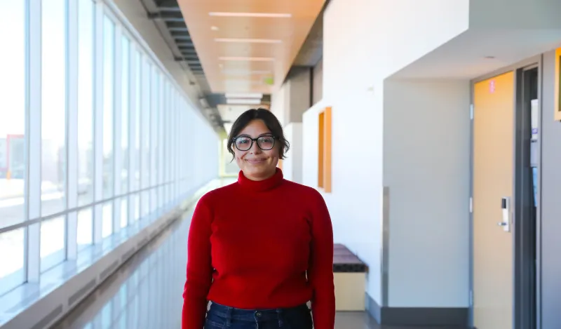 this is a photo of a female NHCC student smiling in the upstairs hallway of the BHCC building. She is wearing a red turtleneck sweater and dark wash jeans with her hair pulled back. 