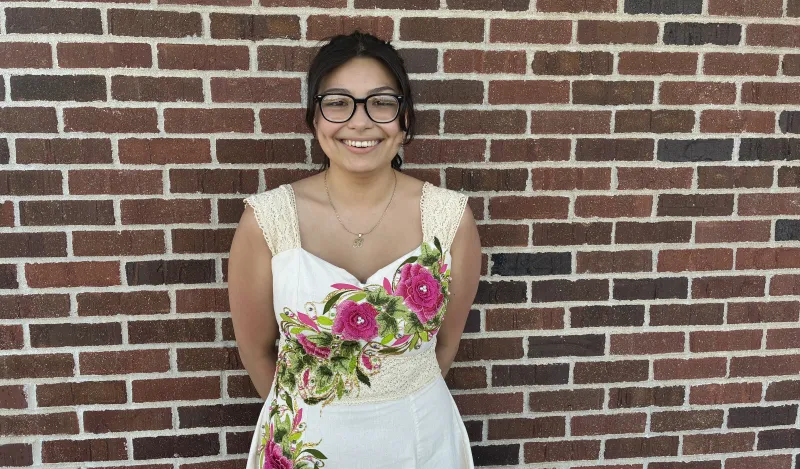 this is a photo of female, NHCC math student, Brenda. She is standing outside, againist a brick wall, wearing a white dress, with handpainted pink roses on it from Mexico. 