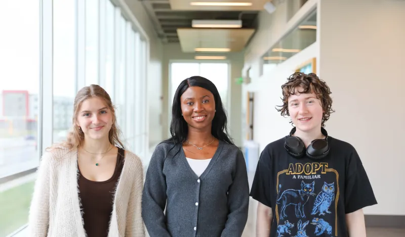 this is a photo of three NHCC students standing side-by-side in the upstairs hallway of the BHCC builidng. Two female students, Anas and Danista stand next to male student, Kenny, posing for a photo and smiling. 