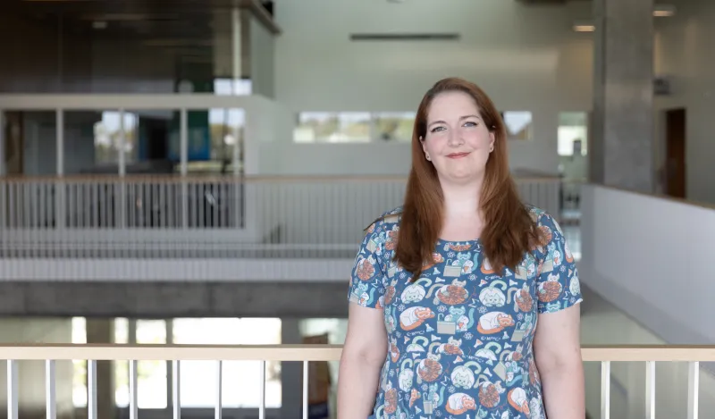 this is a photo of female NHCC student, Jenny Victory, standing in the upstairs hall of the BHCC building, smiling, wearing a short sleeved dress with science illustrations on it. 
