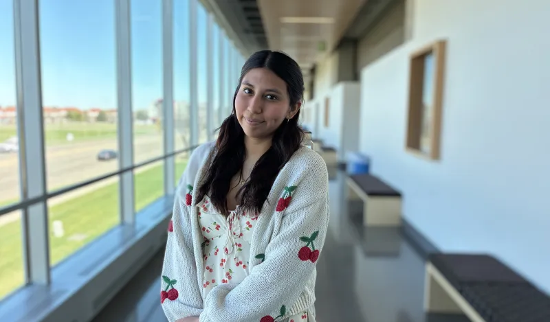 this is a photo of female NHCC student, Reyna Hernandez, standing in the upstairs hall of the BHCC building, smiling, wearing a tank top and a knit cardigan sweater that both have red cherries on them. 