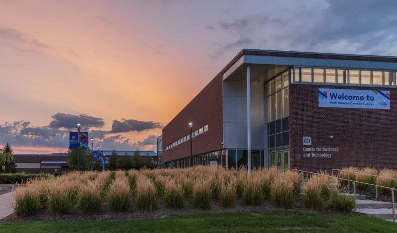 this is a photo taken of NHCC's Center for Business and Technology (CBT) building, outside, facing the side of the building, Door 12 in sight and behind the building is a beautiful light orange and pink, blue and purple-y sunset backdrop. 