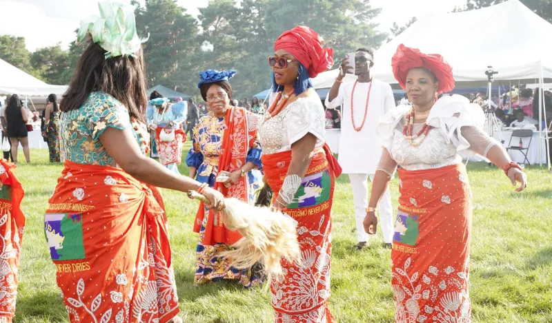 Several attendees wearing vibrant red, white, and multicolored traditional Igbo clothing and headwraps dance in a grassy outdoor festival area during the IgboFest Cultural Celebration. White event tents and other attendees are visible in the background.