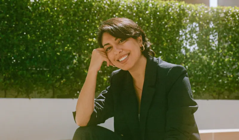 Person with dark hair sitting outside smiling on a sunny day. 