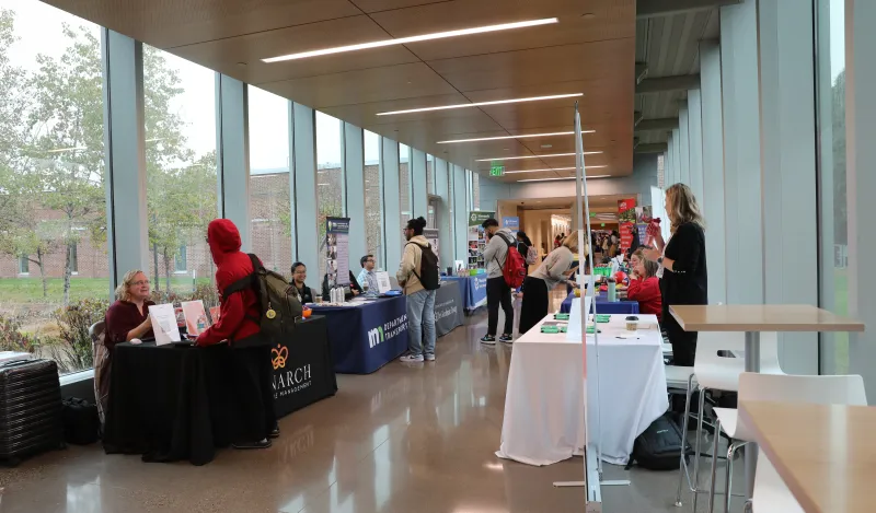 this is a photo of the BHCC hallway during a Career Fair with tables set up on either side of the hall, with students walking and stopping to ask questions to the employers sitting behind the tables.