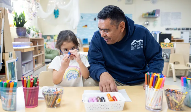 this is a photo of a little girl holding up a blue marker to her face, in the midst of coloring at a table with her dad smiling and sitting next to her.