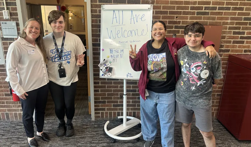 this is a photo of two female NHCC Diversity Specialists standing and smiling with two NHCC students in front of the Gerry Huerth Center in the Campus Center. The whiteboard sign in between them reads, All Are Welcome! 